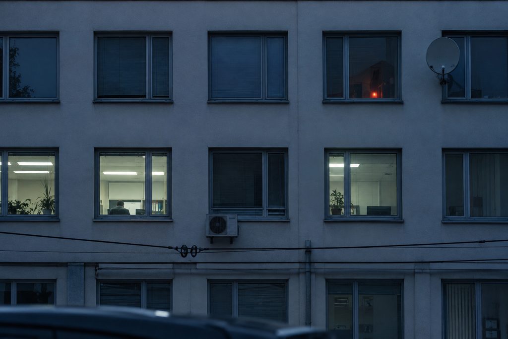 A second-floor office seen from the doorway on a grey February afternoon: three desks, two of them occupied, an uncurtained window.